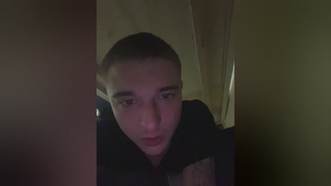 A dimly-lit video of a young boy with short hair, looking up with a neutral expression, wearing a dark jacket. The background is shadowy and indistinct.