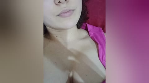 A close-up video of a woman's partially exposed chest, showing medium-sized breasts with a pink bra visible. She has light skin, straight nose, and lips with light pink lipstick. The background is blurred with a gradient of pink and purple hues.