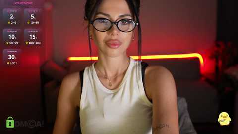 Video of a woman with fair skin, wearing black-rimmed glasses, a white sleeveless top, and a black tattoo on her forearm, seated indoors with a red-lit background, displaying a digital game interface.