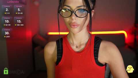 Video of a young woman with fair skin, large black-rimmed glasses, and a red tank top, standing in a dimly lit gym with red lighting and exercise equipment in the background.