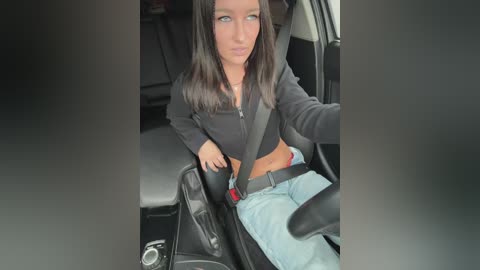 A video of a young woman with long dark hair, wearing a black zip-up hoodie, light blue jeans, and a seatbelt, seated in a car with a grey interior.