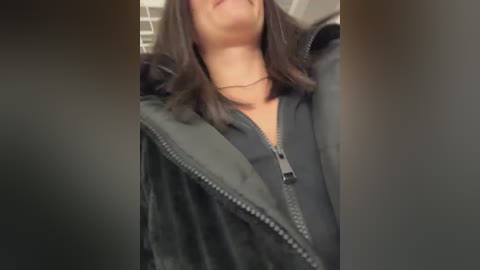 Video of a person with shoulder-length dark hair, wearing a black zip-up jacket, partially unzipped, revealing a black top underneath. Background is blurry with light brown tones.