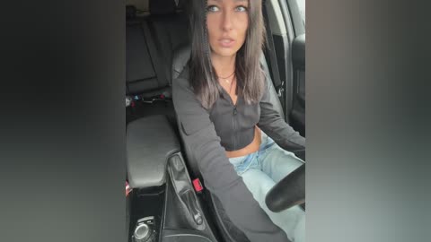 Video of a woman with medium skin tone and straight black hair, wearing a black crop top and light blue jeans, sitting in a car.