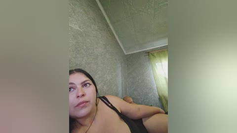 A video shows a Latina woman with medium skin tone, dark hair, and a neutral expression, lying on a bed. She's wearing a black bra, with textured gray walls and a green curtain in the background.