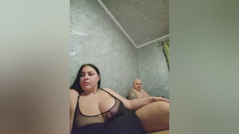 Video of a plus-size woman with large breasts in a sheer black lingerie top, lying on a bed with a bald man, both in a dimly lit room with textured grey walls.