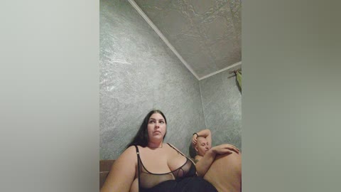 A video of a plus-sized woman with dark hair and a large breast in a sheer black bra, sitting on a bed with a man's hand on her shoulder. The background features textured, light gray walls.