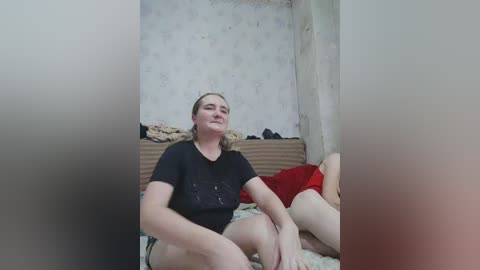 A video of a middle-aged, fair-skinned woman with blonde hair, wearing a black shirt and beige shorts, sitting on a bed with a red pillow and beige blanket, in a dimly lit, worn-out room with floral wallpaper.