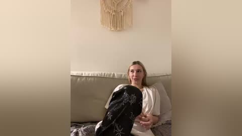 A video of a blonde woman in a beige dress, reclining on a beige couch, with a beige macram\u00e9 wall hanging behind her.