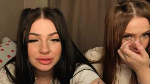 Video of two young women with long black hair, one in pigtails and the other with pigtails, both with fair skin, smiling, one holding a stuffed animal, against a neutral background.