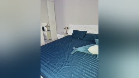 Video of a minimalist bedroom with a white headboard, dark blue striped bedspread, pillows, and a small white bowl. The walls are light gray, and a white dresser is visible in the background.