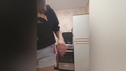 A video of a woman in a black hoodie and ripped shorts, walking away from a kitchen with a white refrigerator, beige wallpaper, and a microwave.