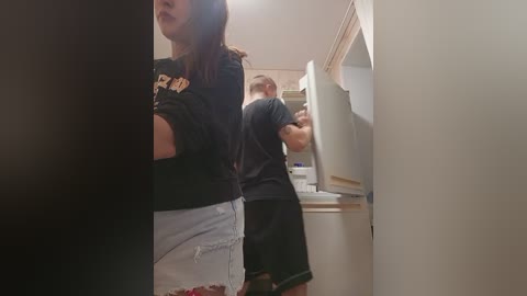 A video captures a woman in a black shirt and white shorts, standing in a kitchen with a man wearing a black shirt, opening the refrigerator.