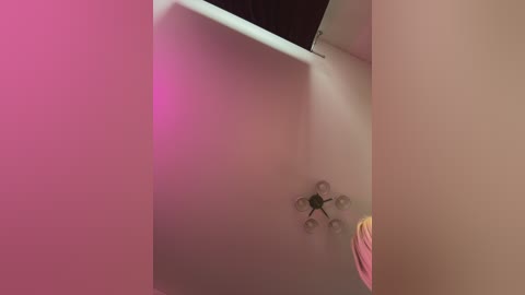 A video of a minimalist, modern bathroom with a beige wall, a pink towel hanging, and a quadcopter drone hovering near the ceiling, bathed in soft pink light.