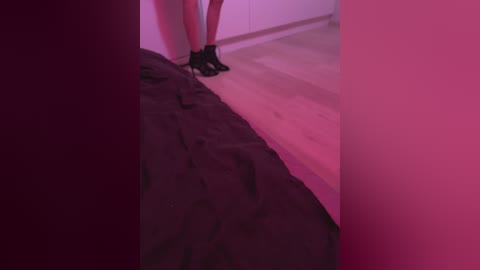 Video: A person in black socks stands on a dark floor, with a bright pink wall and a wooden floor in the background.