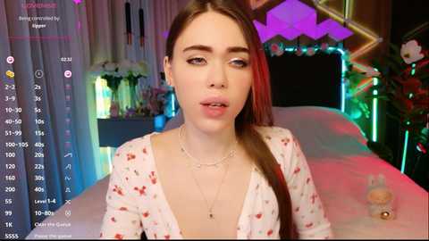 Video of a young woman with light skin and long brown hair, wearing a low-cut floral top, in a colorful, dimly lit bedroom with a bed and floral decor.