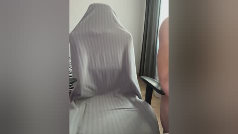 A video shows a man's nude backside sitting in a chair, covered with a light grey blanket. The room features a dark curtain and a wooden floor.