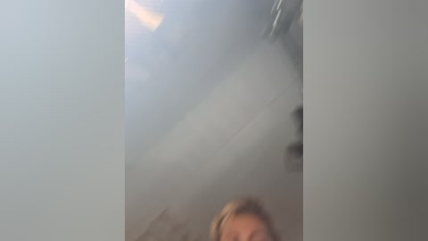 Video of a woman with light skin and blonde hair, partially obscured by fog, standing in a dimly lit room with white walls and a light wooden floor.