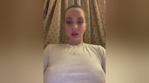 Video of a fair-skinned, young woman with light brown hair, wearing a tight, beige t-shirt that accentuates her large breasts, lying on a beige bed with patterned curtains in the background.
