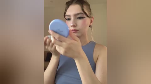 Video of a young woman with fair skin and brown hair in a ponytail, wearing a sleeveless light blue top. She is applying makeup with a blue compact mirror, against a beige wall.