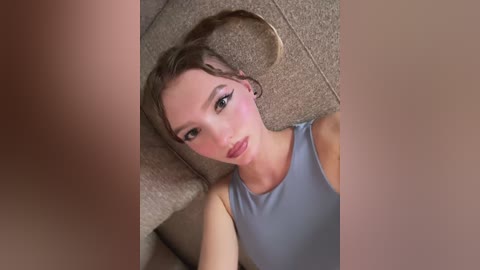 Video of a young woman with light skin and straight brown hair lying on a beige carpet, wearing a gray tank top. Her expression is neutral, with subtle makeup, including eyeliner and mascara.