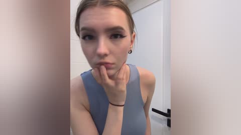 Video of a young woman with light skin and brown hair, wearing a light blue sleeveless top, posing with her hand near her mouth, in a white-tiled bathroom with a black towel rack in the background.