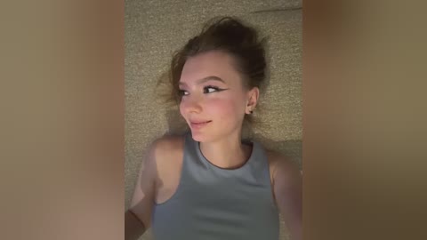 Video of a fair-skinned woman with auburn hair, lying on a beige carpet, wearing a sleeveless gray top, smiling.