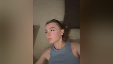 Video of a young Caucasian woman with fair skin, brown hair tied back, wearing a light blue tank top, lying on a beige couch.