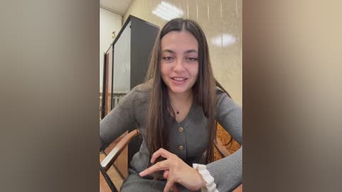 Video of a young woman with long, straight brown hair, wearing a gray cardigan and light-colored shirt, sitting in a wooden chair with a neutral expression, in a room with beige walls and a black door.