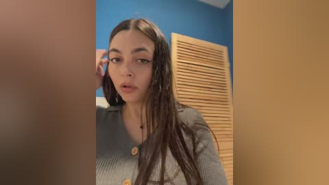 A video of a young woman with long brown hair, fair skin, and a neutral expression, wearing a gray cardigan, standing in a blue-walled room with a wooden shutter background.