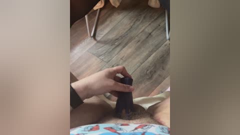 Video of a woman with light skin, wearing a patterned blue shirt, using a black dildo on her shaved vulva, on a wooden floor.