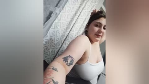 Video of a plus-size woman with medium skin tone, brown hair, and tattoos on her arm. She's wearing a white tank top, sitting in a room with patterned curtains and a grey wall.