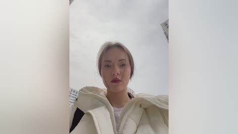 A video of a young woman with light skin and straight, light brown hair, wearing a cream-colored hoodie, standing in a dimly lit room with white walls.