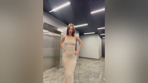 Video of a woman with long, light brown hair, wearing a strapless, form-fitting, nude-colored dress, walking in a dimly lit, modern corridor with dark, textured ceiling panels and bright fluorescent lights.