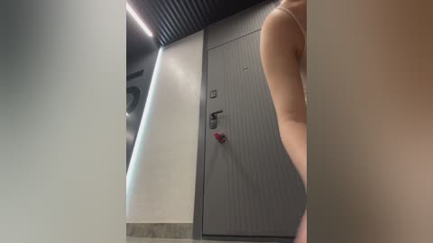 Video of a modern hallway with a gray door, a black wall, and a person's arm and torso partially visible in the right foreground. The setting is clean and minimalist.