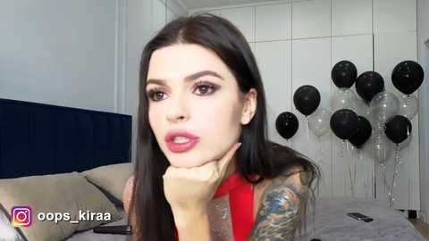 Video of a pale-skinned woman with long, straight black hair, wearing a red dress, leaning on her hand, with a tattooed arm, in a modern bedroom with a bed, black balloons, and white wall.