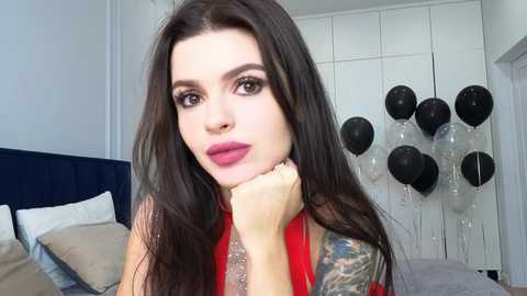Video of a woman with long, dark hair, wearing red lipstick and a red dress, leaning on her hand, in a bedroom with white walls, a navy-blue headboard, and black and silver balloons.