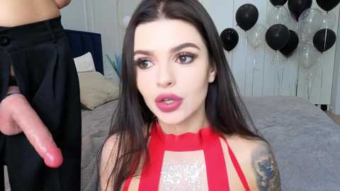 Video of a light-skinned woman with long black hair and pink lipstick, wearing a red halter top, sitting on a bed with black balloons in the background, looking at a man's erect penis.