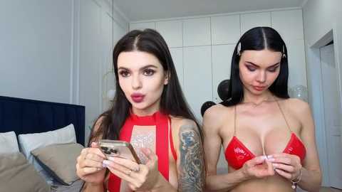 Video of two young women with fair skin and long dark hair. One wears a red halter top, holding a smartphone, and the other a red bikini top, adjusting straps. Both are in a modern, minimalistic bedroom with white walls and a navy-blue upholstered bed.