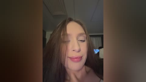 Video of a young woman with long, straight brown hair, fair skin, and closed eyes, smiling slightly. She wears a black top. Background shows a dimly lit room with a ceiling fan.