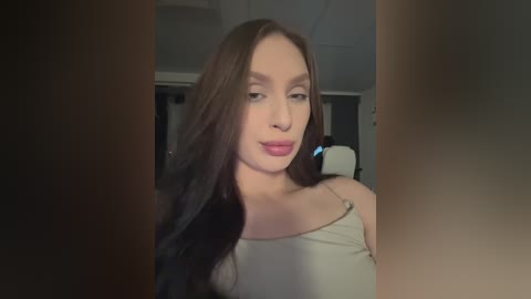 Video of a young woman with long brown hair, light skin, and red lipstick, wearing a beige tank top. She's indoors, with a blurred background featuring a chair and dim lighting.
