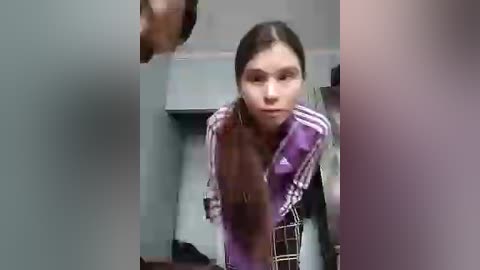 A blurred video of a young woman with straight brown hair, wearing a purple Adidas hoodie, crouching near a bed in a dimly lit room.