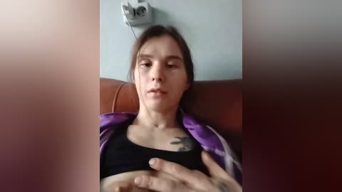 Video of a pale-skinned, androgynous person with brown hair, wearing a purple jacket and black bra, reclining on a brown leather couch, with a power outlet in the background.