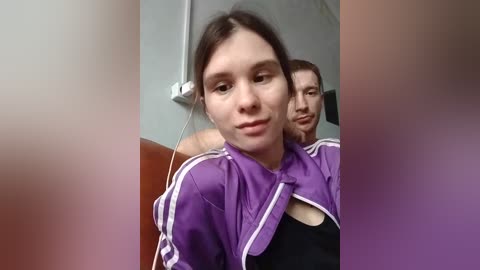 Video of two people: a young woman with light skin, brown hair, wearing a purple jacket, and a man with short brown hair, both in a public restroom.