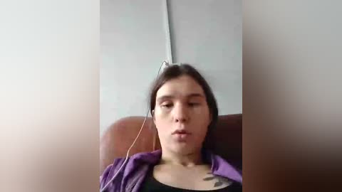 Video of a young woman with straight, dark hair, wearing a purple jacket, lying on a brown couch with headphones on, in a dimly lit room with white walls.