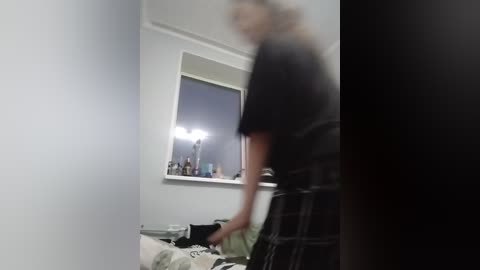 A blurred video of a person in a black t-shirt and plaid pants standing near a bed, with a bright light reflecting off a window behind them.