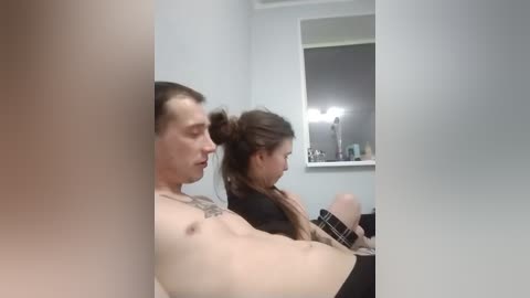 Video of a muscular shirtless man with a tattoo, sitting next to a woman with long brown hair in a ponytail, both facing a mirror in a dimly lit bathroom.