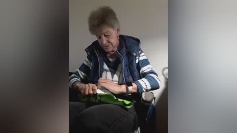 A video of an elderly white woman with short gray hair, wearing a blue and white striped sweater and a puffy vest, seated in a wheelchair, fiddling with a green object, in a dimly lit room.