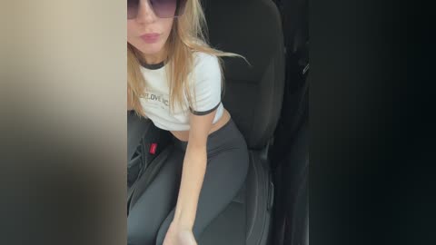 Video of a blonde woman with fair skin, wearing a white crop top and black leggings, sitting in a car, partially obscured by a window.