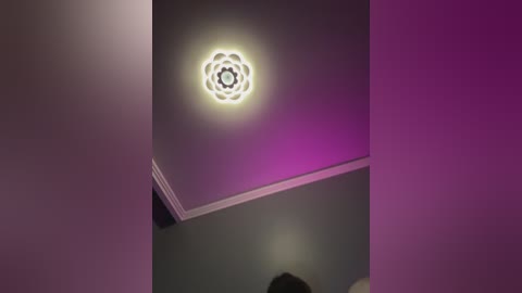 Video of a modern ceiling with a circular, glowing, flower-like light fixture in the center, surrounded by purple and pink lighting, set against a dark gray wall.