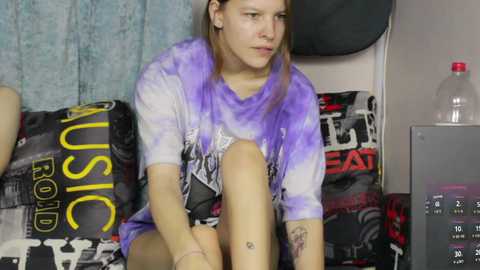 Video of a young woman with long brown hair, wearing a tie-dye shirt and black shorts, sitting on a bed with graffiti-themed pillows, looking introspective.